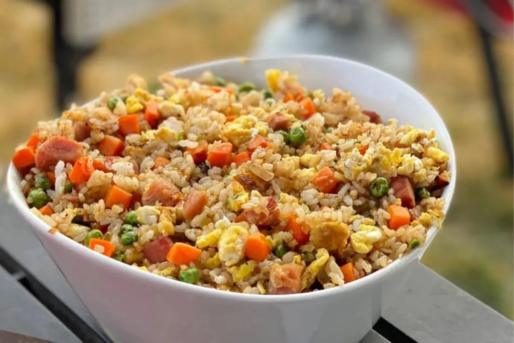 A white bowl filled with Budget Friendly Rice Meals like fried rice mixed with scrambled eggs, diced ham, peas, and carrots, placed on an outdoor table.